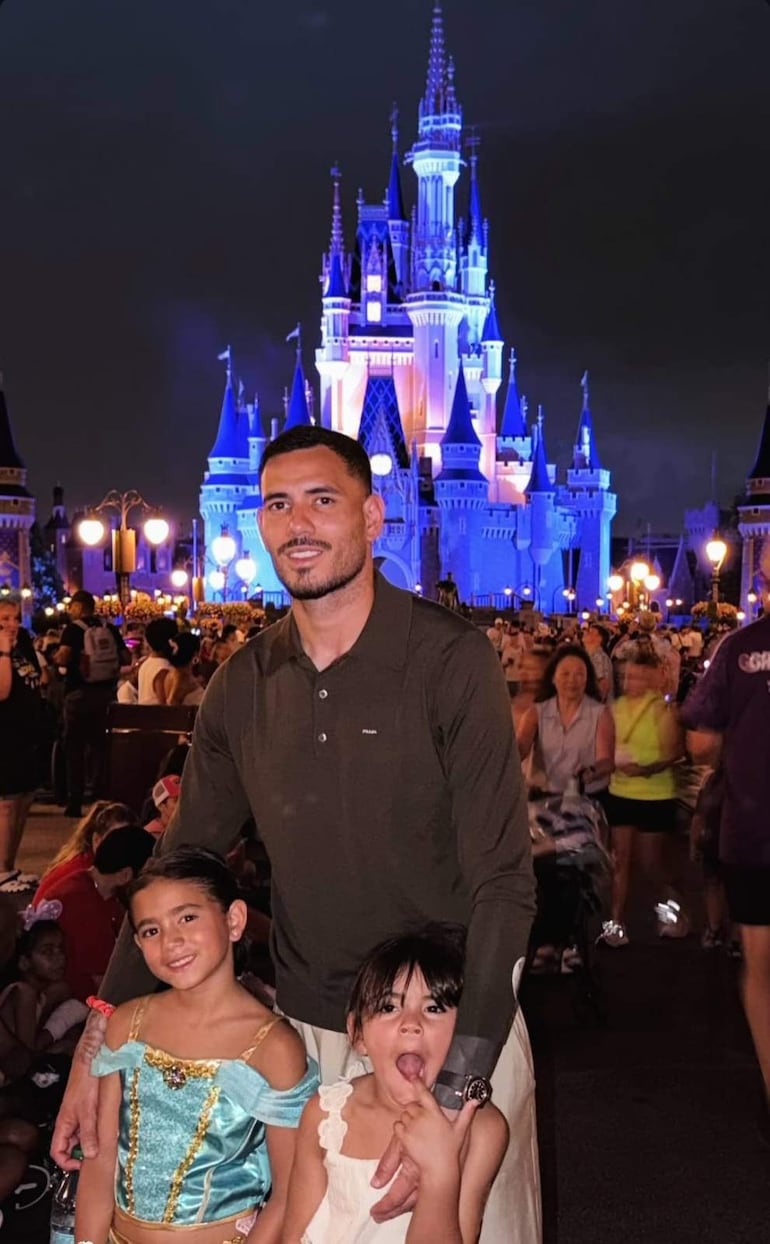 Antonio Sanabria sonríe, sosteniendo a una niña vestida como princesa, mientras otra sonríe y hace gestos divertidos, con el castillo iluminado al fondo.