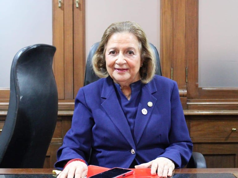 Alicia Pucheta, mujer de cabello rubio y rizado, viste traje azul, sonríe sentada en su oficina con documentos y un teléfono móvil.