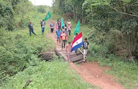 Grupo de personas marchando con banderas, rodeados de vegetación, dos hombres observan desde el primer plano.