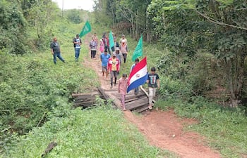 Grupo de personas marchando con banderas, rodeados de vegetación, dos hombres observan desde el primer plano.