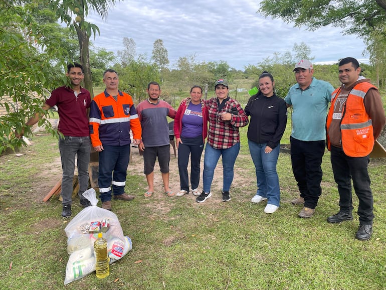 Ocho personas sonrientes, algunas con camisetas de colores, sosteniendo bolsas con víveres en un entorno rural.