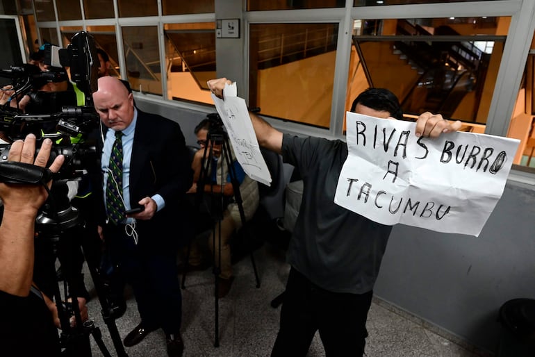 Hombre con camiseta gris sostiene un cartel en mano, rodeado de periodistas en un ambiente interior del Palacio de Justicia.