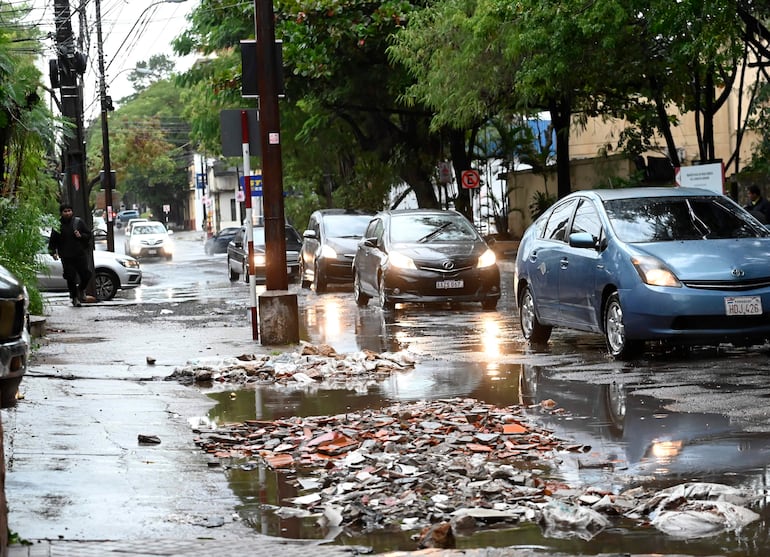 Calle urbana con pavimento mojado y vehículos en movimiento, rodeada de vegetación tras la lluvia.