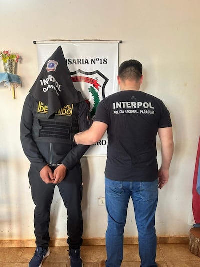 Hombre con pasamontañas y manos esposadas, custodiado por agente de INTERPOL en ambiente interior de oficina.