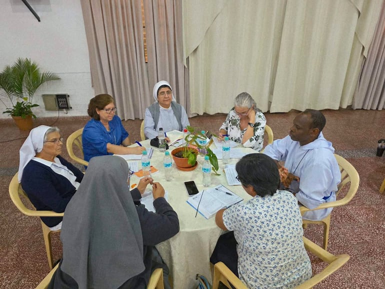 Siete personas, tres con hábitos religiosos, conversan a la mesa, en un ambiente claro y formal con plantas y material de oficina.