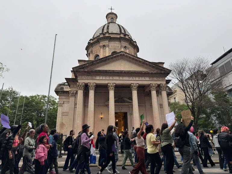 Grupo de mujeres marchando con pancartas en una calle nublada, algunas sosteniendo la bandera nacional.