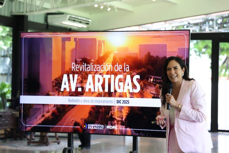 Claudia Centurión sonríe mientras sostiene un micrófono, rodeada de un ambiente iluminado con una pantalla de presentación al fondo.