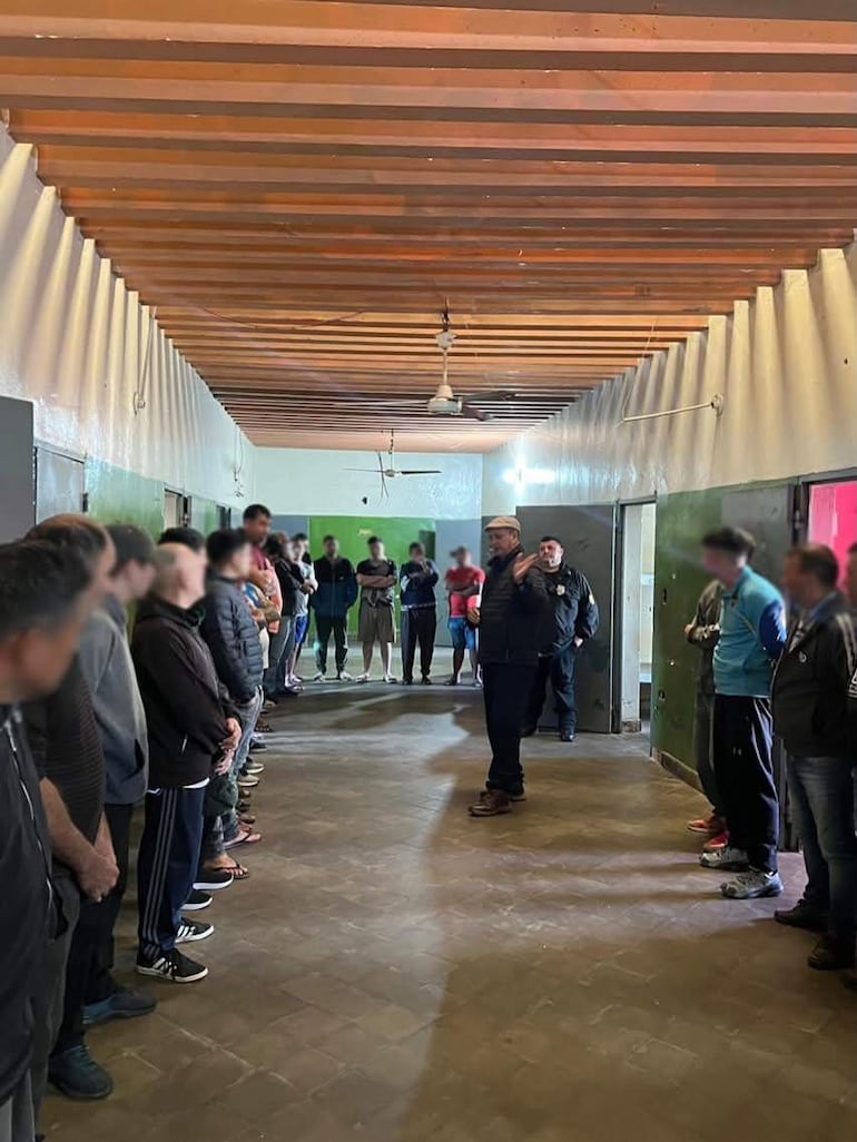Hombre de pie con ropa casual habla a un grupo de 31 personas en una habitación con paredes verdes y grises.