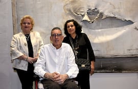 Félix Toranzos en silla de ruedas, con camisa blanca, entre dos mujeres, una de blusa clara y otra de negro, en un ambiente artístico.