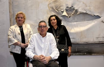 Félix Toranzos en silla de ruedas, con camisa blanca, entre dos mujeres, una de blusa clara y otra de negro, en un ambiente artístico.