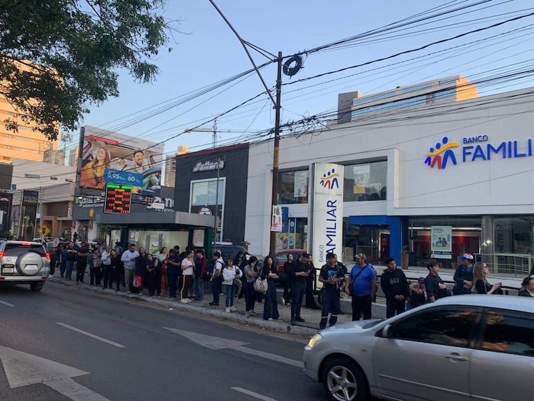 Fila de personas de pie y sentadas, algunas con teléfonos móviles, frente al Banco Familiar en un ambiente urbano diurno.