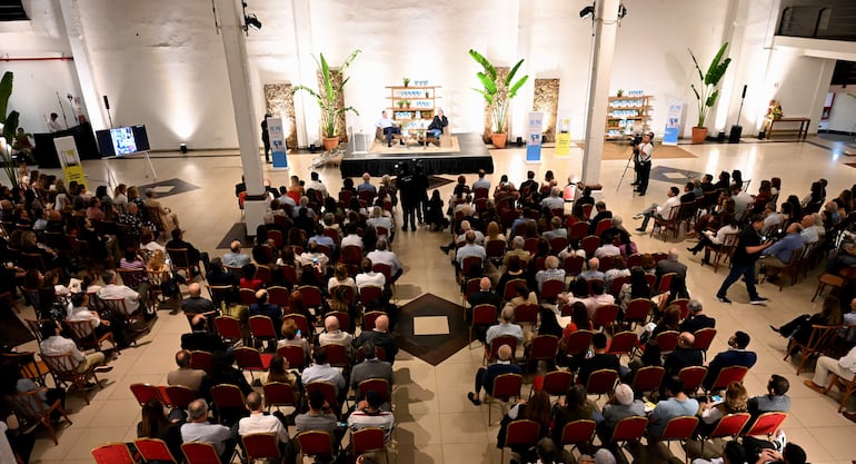 Jorge Mendelzon en mesa de presentación, rodeado de asistentes atentos en un espacio elegante con sillas rojas y plantas decorativas.