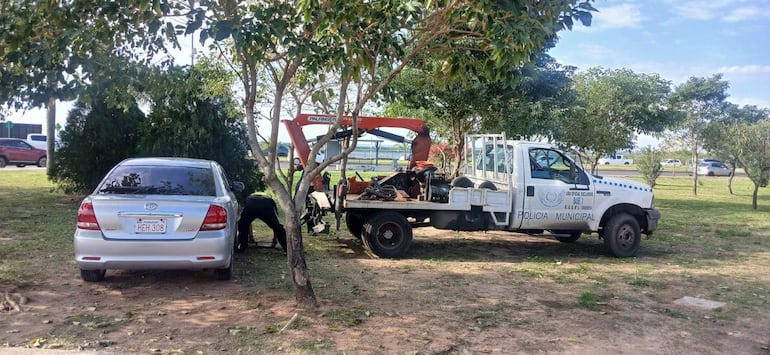 Vehículo de la policía con grúa amarilla y operario agachado cerca de un automóvil plateado estacionado, rodeado de árboles.