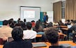 Hombre en blazer negro y camisa azul presenta a jóvenes economistas en aula con escritorios y laptops abiertas.