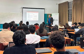 Hombre en blazer negro y camisa azul presenta a jóvenes economistas en aula con escritorios y laptops abiertas.