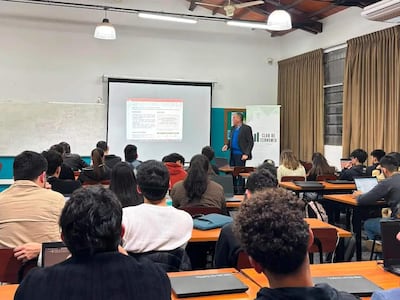 Hombre en blazer negro y camisa azul presenta a jóvenes economistas en aula con escritorios y laptops abiertas.