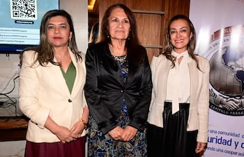 Tres mujeres sonríen en un evento formal; una lleva chaqueta blanca, otra vestido oscuro y una más, camisa blanca con falda negra.