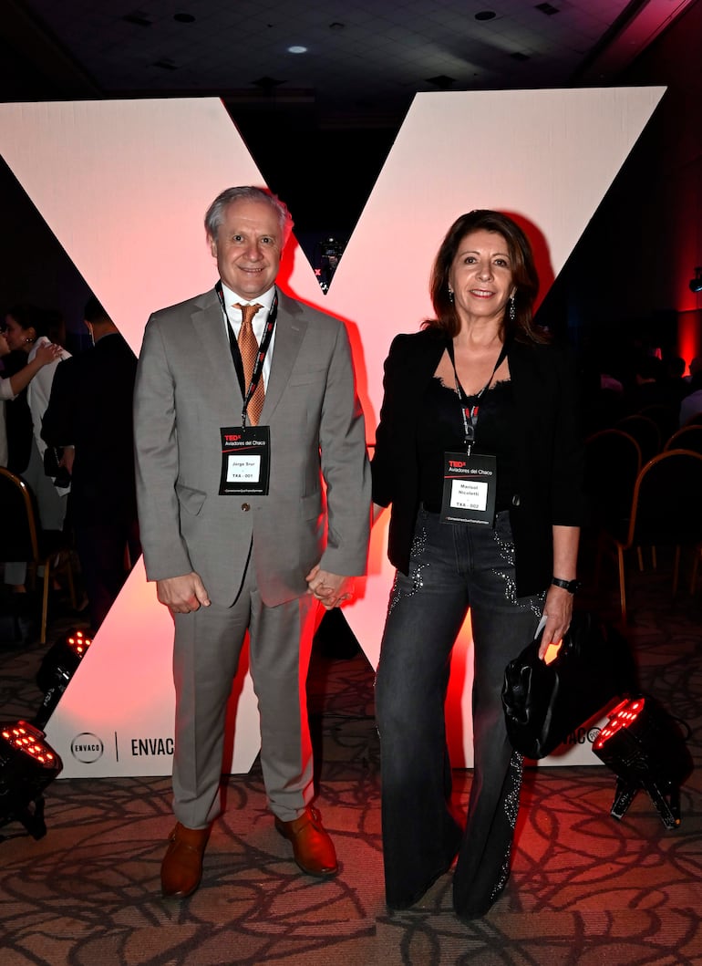 Jorge Srur en traje claro y Marisol Nicoletti en top negro, sonriendo frente a un panel iluminado del evento TEDx.