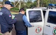 Un policía guía a Senón Caceres Duarte, vestido con chaleco gris y camisa clara, hacia una camioneta policial.