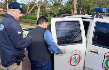 Un policía guía a Senón Caceres Duarte, vestido con chaleco gris y camisa clara, hacia una camioneta policial.