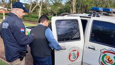 Un policía guía a Senón Caceres Duarte, vestido con chaleco gris y camisa clara, hacia una camioneta policial.