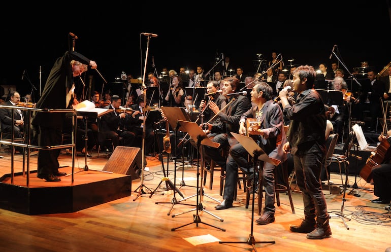 Director de orquesta con traje negro dirige a Los Nocheros en una actuación en un escenario iluminado con coloridas luces.