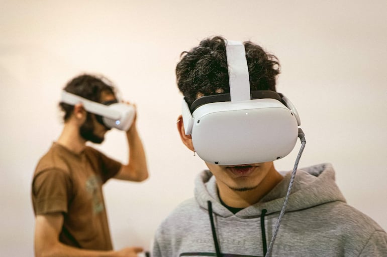 Joven con máscara de realidad virtual blanca y sudadera gris interactúa con tecnología, mientras otro joven con cabello rizado está en el fondo.