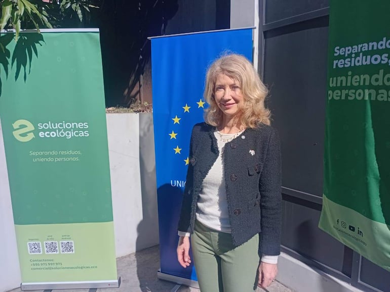 Mujer de cabello rubio y ondulado lleva abrigo negro y pantalones verdes, sonriendo frente a banners alusivos a soluciones ecológicas.