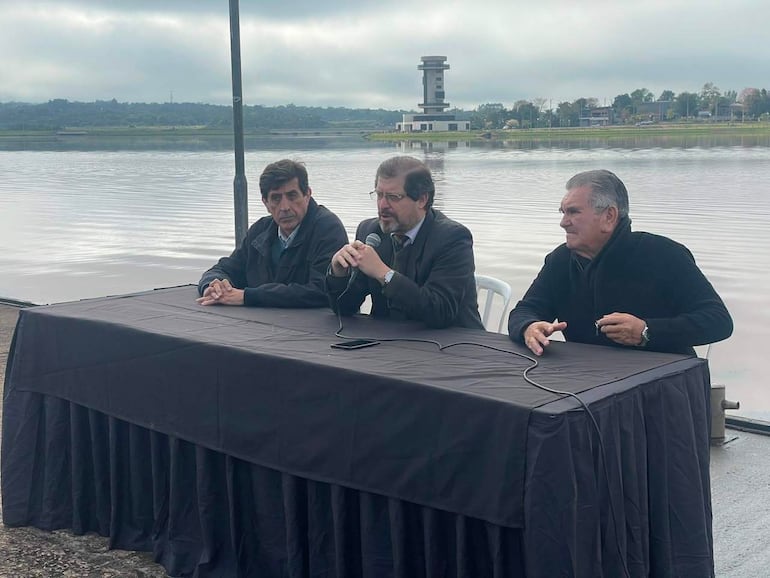 Tres hombres sentados tras mesa negra; uno habla con micrófono, rodeados de vegetación y un cuerpo de agua de fondo.
