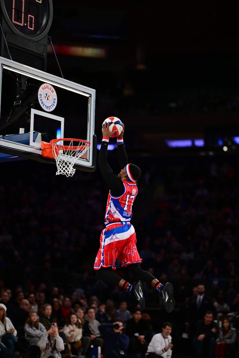 Atleta del equipo Harlem Globetrotters en uniforme rojo y azul, realizando una espectacular volcadura en la cancha, rodeado de un público entusiasta.