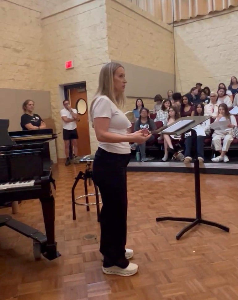 Mujer con cabello lacio y ropa blanca gesticula mientras habla frente a un piano y un grupo de personas observan.