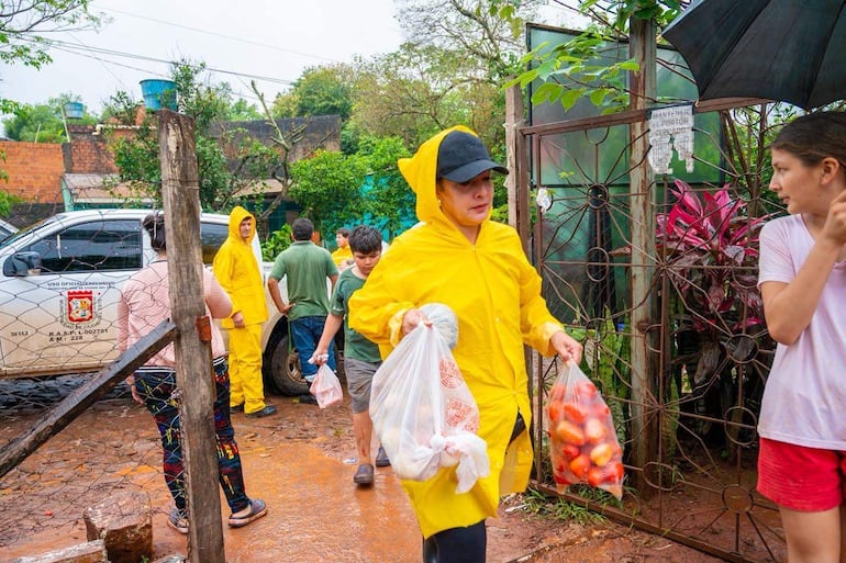 Mujer con abrigo amarillo y niña en camiseta blanca sosteniendo bolsas, rodeadas de personas en un ambiente lluvioso.