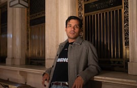 El David Aguilar con camiseta negra y abrigo de cuadros, sonríe en una estación con detalles arquitectónicos elegantes.