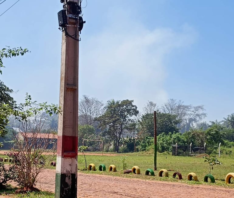 Poste de electricidad con vegetación y neumáticos de colores, bajo un cielo azul y humo en el aire.