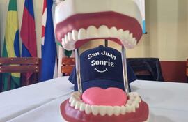 Modelo de boca abierta con dientes artificiales sobre mesa blanca, gorra azul con 'San Juan Sonrie' y banderas al fondo.
