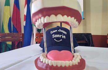 Modelo de boca abierta con dientes artificiales sobre mesa blanca, gorra azul con 'San Juan Sonrie' y banderas al fondo.