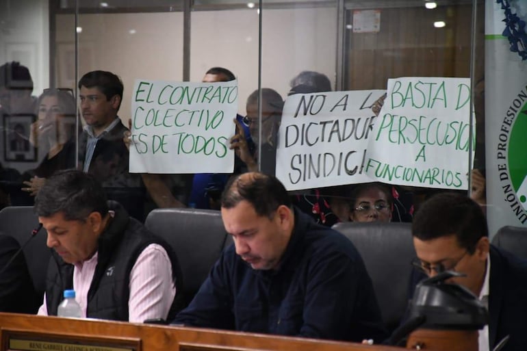 Cuatro hombres concentrados en documentos, mientras en el fondo varias personas sostienen carteles de protesta contra la dictadura sindical.