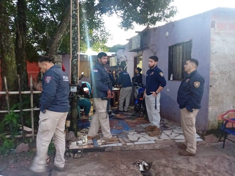 Seis hombres policías en chaquetas oscuras frente a vivienda morada, un agente trabaja en un motor entre vegetación.