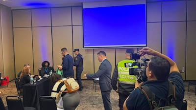 Mujer de blusa verde habla con un hombre, mientras dos agentes de seguridad observan en el fondo, en un ambiente profesional.
