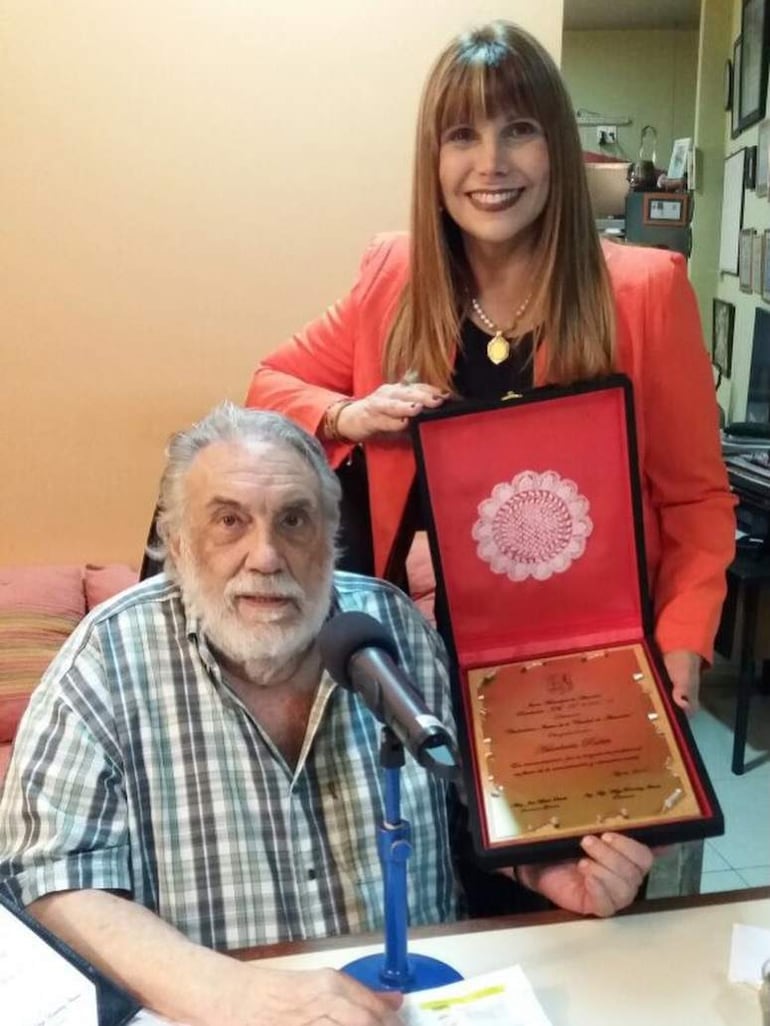 Pelusa Rubin con barba canosa sentado frente a un micrófono, mientras una mujer con blazer naranja sostiene un premio detrás.