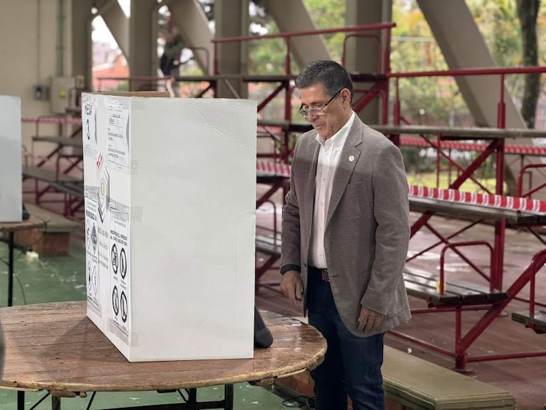 Horacio Cartes, vestido con saco gris y camisa blanca, observa la urna electoral en un recinto público.