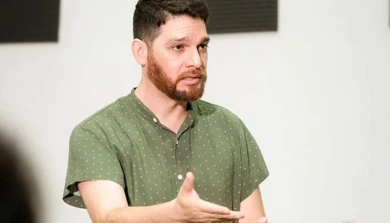 Héctor Lozzca de pie, gesticulando con la mano derecha en un entorno interior con paneles oscuros de fondo.