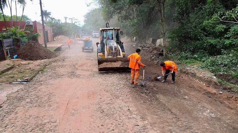 Cuatro hombres en uniformes naranjas trabajando con pala y azada en una calle polvorienta, junto a una retroexcavadora.