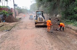 Cuatro hombres en uniformes naranjas trabajando con pala y azada en una calle polvorienta, junto a una retroexcavadora.