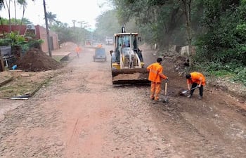 Cuatro hombres en uniformes naranjas trabajando con pala y azada en una calle polvorienta, junto a una retroexcavadora.