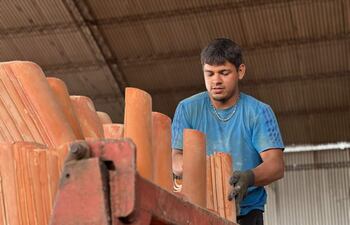 Hombre en camiseta azul y guantes oscuros organiza tejas terracota apiladas en un vehículo en un ambiente industrial.