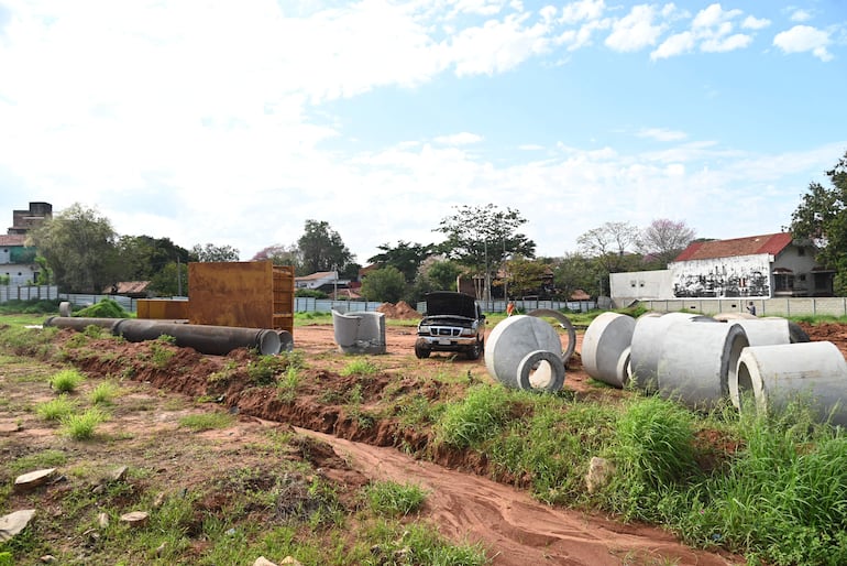 Terreno con tubos de concreto, un contenedor metálico y un coche en mantenimiento bajo un cielo parcialmente nublado.