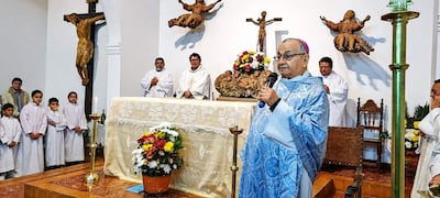 Monseñor Medina, vestido de sacerdote azul, habla con tres hombres en toga blanca en un entorno religioso con estatuas y flores.