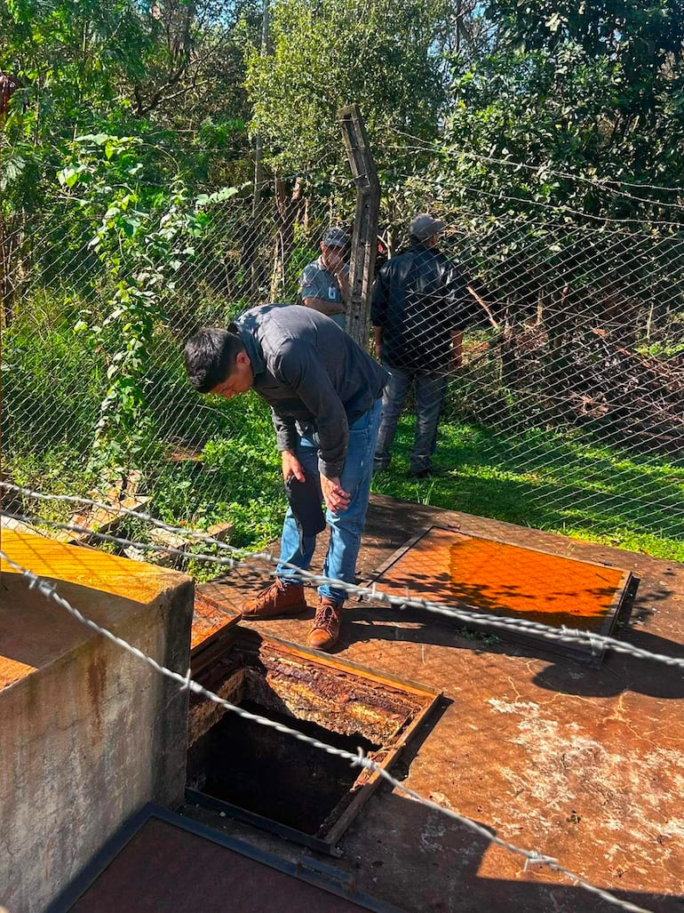 Hombre en camisa de mezclilla inspecciona acceso subterráneo, acompañado de tres hombres en ropa casual en un entorno rural.