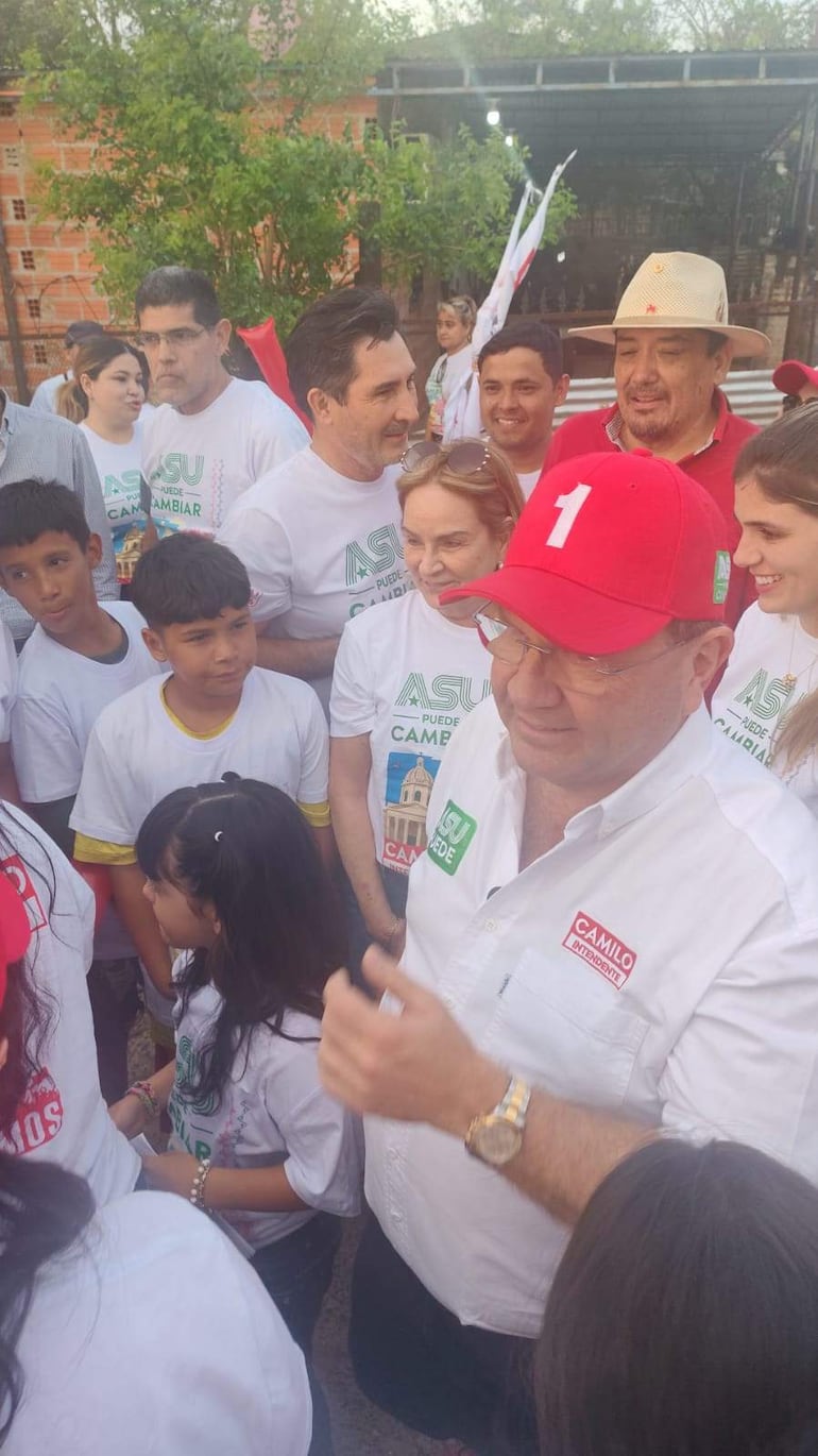 Camilo Pérez con gorra roja y camiseta blanca rodeado de personas en ambiente exterior, interactuando y sonriendo.
